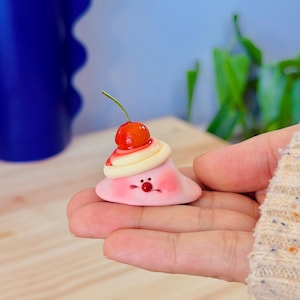 May include: A whimsical, pink, cloud-shaped figurine with a cherry on top. The figurine has a smiling face with rosy cheeks and a red nose. The cherry is bright red with a green stem, and the figurine has a white and red swirl design.