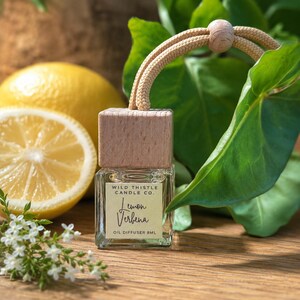 May include: A small square glass oil diffuser bottle with a light wood cap and a beige hanging cord. The label reads Wild Thistle Candle Co. Lemon Verbena Oil Diffuser 8ml. The bottle is positioned near a sliced lemon and green leaves.