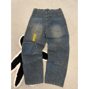 May include: Blue denim jeans featuring a patchwork design with multiple panels of varying blue shades. The trousers include two back pockets, a rectangular leather-like patch on the waistband, and a small, bright yellow rectangular tag on the left leg.