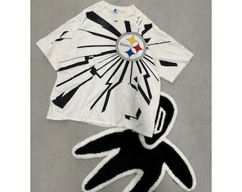 Puede incluir: Camiseta blanca con un diseño gráfico negro y el logotipo de los Steelers. En la imagen también hay una alfombra en blanco y negro con una forma única. La camiseta tiene cuello redondo y mangas cortas.