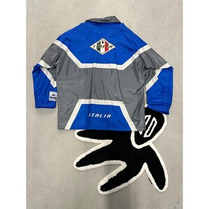 May include: A blue, gray, and white Italia jacket with a diamond-shaped emblem and the word "ITALIA" on the back. The jacket is laid on a black and white rug.