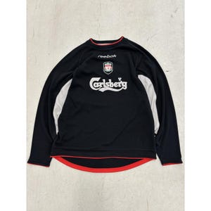 May include: A black long-sleeve athletic shirt featuring the Reebok logo, a Liverpool Football Club crest, and the Carlsberg brand name in white text. The shirt includes grey side panels and red trim along the collar and the bottom hemline.
