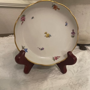 May include: A white porcelain saucer with a scalloped edge and a gold-painted rim. The surface features small, hand-painted floral motifs in various colors, including pink, yellow, blue, and purple, scattered across the center and the border.