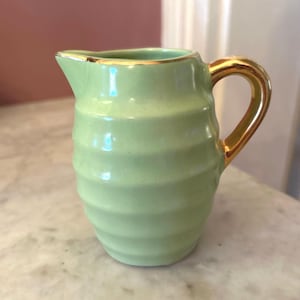 May include: A small, light green ceramic creamer with a gold handle and rim. The creamer has a ribbed design around the body. The handle is a curved, gold color.