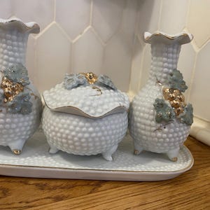 Op de afbeelding: Een witte porseleinen toilettafelset met een gestructureerd, gestippeld oppervlak. De set bevat twee vazen en een pot met deksel, allemaal versierd met gouden en groene druiventrossen. De stukken staan op een bijpassend ovaal dienblad.