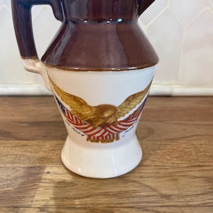 May include: A ceramic pitcher with a glossy brown upper section and a white lower body. The front features a graphic of a golden eagle with outstretched wings draped in an American flag, with the text The Spirit of Liberty printed below.