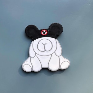 May include: A white cartoon dog-like figure wearing a black hat with Mickey Mouse ears. The hat has a red circle with a white Mickey Mouse icon. The figure is sitting down with its paws in front.