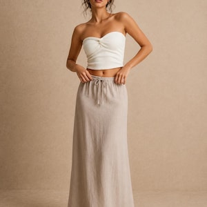 May include: A white strapless tube top with a ribbed texture and a center twist detail paired with a floor-length beige maxi skirt featuring an elasticized waistband and a functional drawstring tie with small tassels at the ends.