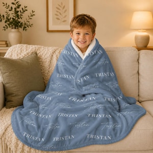 May include: A soft, dusty blue plush blanket featuring a repeating pattern of the name Tristan printed in both a bold serif font and a delicate cursive script. The blanket has a white sherpa-style lining visible along the folded top edge.