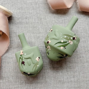 Puede incluir: Dos dreidels verde salvia con dise&ntilde;os florales pintados a mano en blanco, rosa y marr&oacute;n. Los dreidels tienen una forma cl&aacute;sica de cuatro lados y un tallo para girar. El fondo es de tela de lino gris.