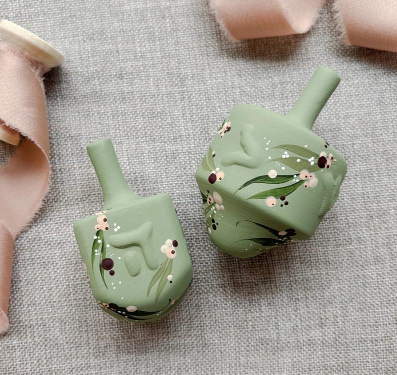 Puede incluir: Dos dreidels verde salvia con dise&ntilde;os florales pintados a mano en blanco, rosa y marr&oacute;n. Los dreidels tienen una forma cl&aacute;sica de cuatro lados y un tallo para girar. El fondo es de tela de lino gris.