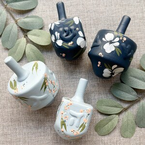 Puede incluir: Cuatro dreidels pintados a mano en tonos azules. Dos son azul marino con dise&ntilde;os florales blancos, y dos son azul claro con acentos melocot&oacute;n y verdes. Los dreidels est&aacute;n dispuestos sobre una superficie de lino neutra con ramitas de hojas verdes.