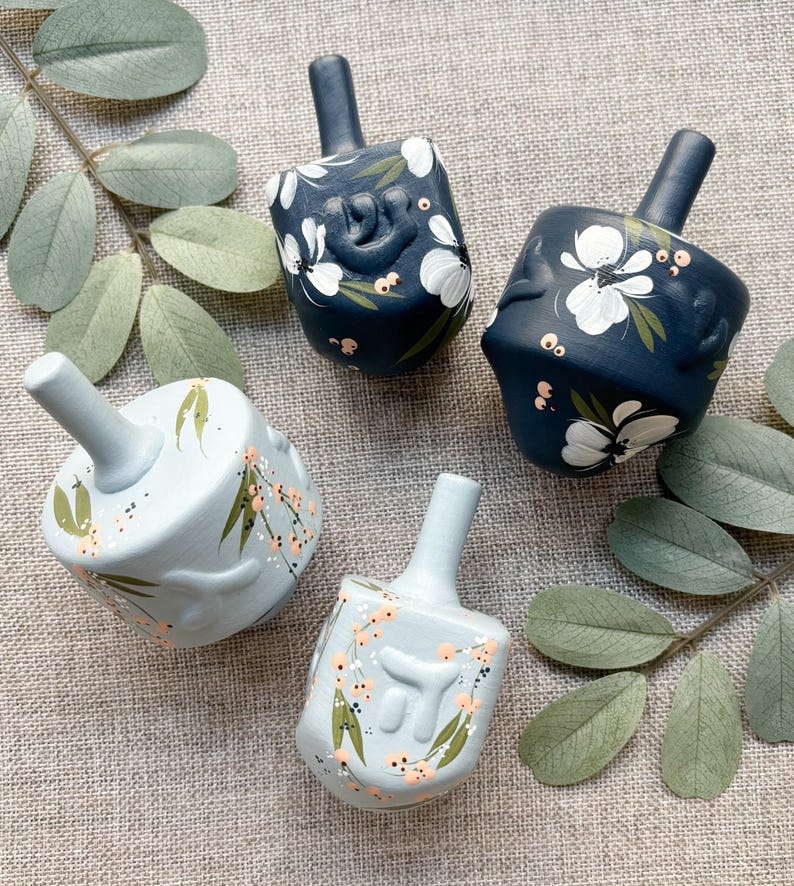 Puede incluir: Cuatro dreidels pintados a mano en tonos azules. Dos son azul marino con dise&ntilde;os florales blancos, y dos son azul claro con acentos melocot&oacute;n y verdes. Los dreidels est&aacute;n dispuestos sobre una superficie de lino neutra con ramitas de hojas verdes.