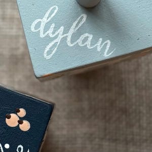 Puede incluir: Dos peque&ntilde;os bloques de madera pintados. Uno es azul claro con el nombre "dylan" en escritura blanca. El otro es azul oscuro con un dise&ntilde;o floral. Los bloques est&aacute;n sobre una superficie de tono neutro.