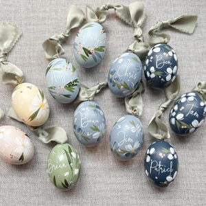 May include: A collection of ten egg-shaped ornaments in various pastel and dark tones, each hand-painted with floral motifs and a unique name in white script. Each ornament features a sage green silk ribbon attached to a gold-tone metal loop.