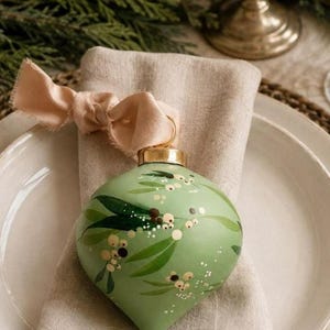 May include: A sage green teardrop-shaped ornament featuring a hand-painted botanical pattern of dark green leaves and small cream-coloured berries. The ornament has a metallic gold cap and is tied with a soft, light peach-coloured fabric ribbon.