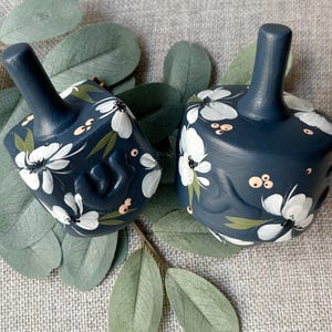Puede incluir: Dos dreidels azul marino con dise&ntilde;os florales blancos y hojas verdes. Los dreidels tienen un tallo en la parte superior y est&aacute;n decorados con flores blancas pintadas a mano, hojas verdes y peque&ntilde;os acentos rosados. Los dreidels est&aacute;n colocados sobre una superficie gris con hojas verdes.