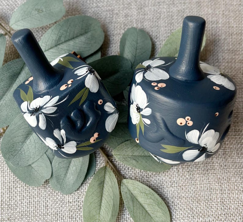 Puede incluir: Dos dreidels azul marino con dise&ntilde;os florales blancos y hojas verdes. Los dreidels tienen un tallo en la parte superior y est&aacute;n decorados con flores blancas pintadas a mano, hojas verdes y peque&ntilde;os acentos rosados. Los dreidels est&aacute;n colocados sobre una superficie gris con hojas verdes.