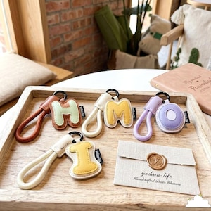 May include: A collection of letter-shaped keychains in various colors, including brown, green, yellow, purple, and white. Each keychain features a different letter and a matching wrist strap. The keychains are displayed on a wooden tray, with a small pouch that reads "yxidian-life Handcrafted Letter Charms."