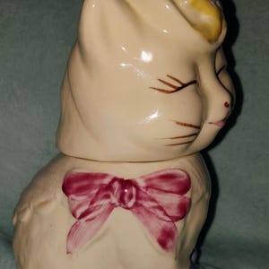 Pode incluir: Estatueta de gato de cerâmica creme com laço rosa e detalhes azuis. O gato usa um chapéu com detalhes amarelos e azuis. O gato está sentado com os olhos fechados.