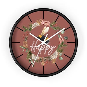 Könnte beinhalten: Eine runde Wanduhr mit schwarzem Rahmen. Das Zifferblatt ist in einem gedämpften Rotbraun gehalten und zeigt einen Blumenkranz, eine Biene und das Wort "Happy" in weißer Schrift. Die Zeiger der Uhr sind weiß.