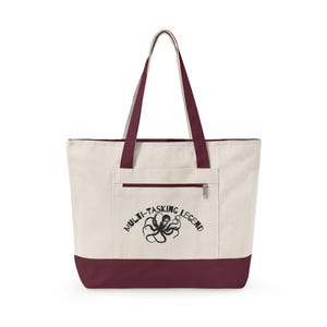 May include: Cream-colored tote bag with a burgundy base, handles, and trim. The bag features a front pocket with a zipper and the text "MULTI-TASKING LEGEND" with an octopus illustration in black.