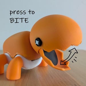May include: An orange toy chick hatching from an egg, with the text "press to BITE". The chick has orange legs and a white diaper. The beak opens to reveal a black interior.