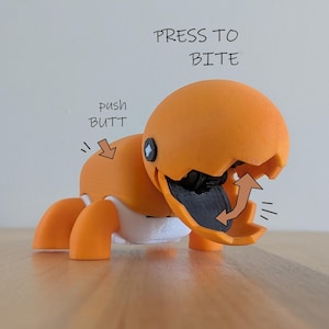 May include: An orange toy creature with a cracked eggshell head and open mouth. The text "PRESS TO BITE" is above the head, and "push BUTT" is on the back. The mouth is black, and the legs are orange.