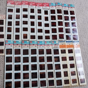May include: A collection of vintage GAF Pana-Vue slide film strips, featuring various travel scenes. The slides are in their original packaging, with red, blue, and orange labels. The image shows a variety of slides, offering a glimpse into the past.