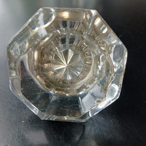 May include: A clear, octagonal glass door knob featuring a faceted, starburst-like pattern radiating from the center. The knob has a smooth, reflective surface with beveled edges, showcasing a transparent design that catches light across its geometric shape.