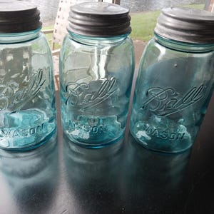 Pode incluir: Três potes de vidro azul-claro em estilo vintage com tampas de rosca de metal prateado. Cada pote apresenta a palavra Ball em relevo em letra cursiva e a palavra MASON em letras de forma na parte frontal, com um formato cilíndrico clássico.