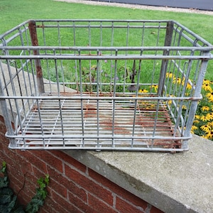 May include: A rectangular wire storage basket constructed from galvanized metal with a grid pattern. The basket features a reinforced top rim and vertical support bars, showing visible signs of oxidation and rust along the base and corners.