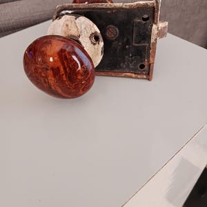 May include: An antique door lock with two oval-shaped, brown and cream-colored knobs. The lock mechanism is black with a white and cream-colored frame, showing signs of wear. The knobs have a glossy, marbled appearance.