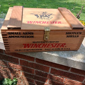 May include: A rectangular wooden storage box with a hinged lid and a brass latch. The top features a black Winchester logo with a rider on horseback. The front is printed with "SMALL ARMS AMMUNITION," "SHOTGUN SHELLS," and "WINCHESTER" in red.