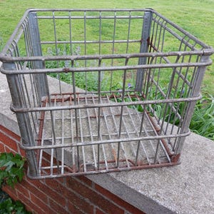 May include: A rectangular, open-top metal wire basket with a grid pattern. The basket has a weathered, gray finish with some rust. It sits on a concrete ledge in front of a brick wall and green grass.