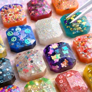 Puede incluir: Una colección de piezas de resina rectangulares y coloridas con purpurina, cuentas y elementos decorativos incrustados. Las piezas presentan varios colores, incluyendo naranja, azul, morado y blanco, con caras sonrientes, mariposas y otros diseños.