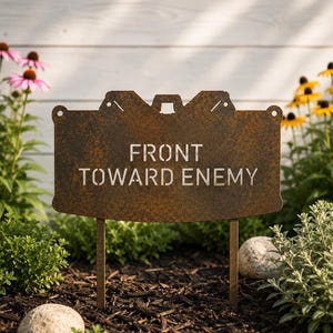 May include: A rectangular metal garden sign with a rusted finish featuring the text FRONT TOWARD ENEMY cut out in a stencil font. The sign has two metal stakes at the bottom for insertion into the ground and circular holes at the top corners.
