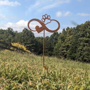 May include: A rusty metal garden stake featuring an infinity symbol with a heart and a paw print. The stake is placed in a grassy area with a wooden fence and trees in the background. The sky is blue with some clouds.