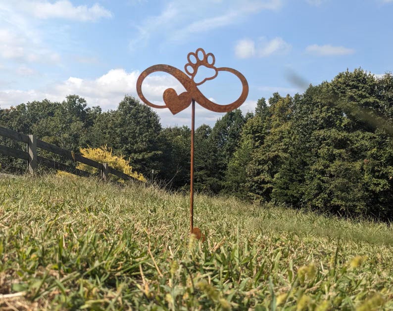 May include: A rusty metal garden stake featuring an infinity symbol with a heart and a paw print. The stake is placed in a grassy area with a wooden fence and trees in the background. The sky is blue with some clouds.