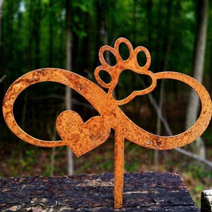 May include: A rusty metal garden stake featuring an infinity symbol, a heart, and a paw print. The stake is designed to be placed in a garden or outdoor setting. The background is a blurred view of a forest.