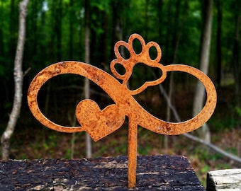 May include: A rusty metal garden stake featuring an infinity symbol, a heart, and a paw print. The stake is designed to be placed in a garden or outdoor setting. The background is a blurred view of a forest.