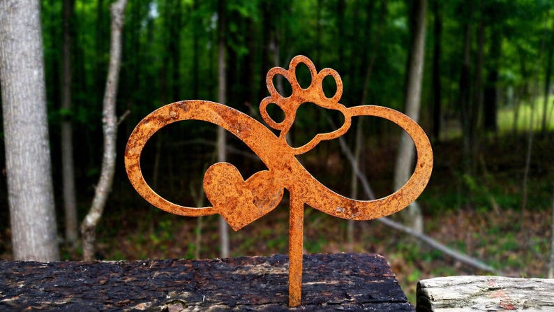 May include: A rusty metal garden stake featuring an infinity symbol, a heart, and a paw print. The stake is designed to be placed in a garden or outdoor setting. The background is a blurred view of a forest.