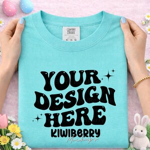 Może przedstawiać: Jasnoniebieska koszulka z napisem "YOUR DESIGN HERE" dużymi, czarnymi literami w stylu retro, ozdobiona gwiazdkami. Nazwa marki "KIWIBERRY Mockups" znajduje się na dole. Koszulka jest trzymana przez dłonie z pomalowanymi paznokciami.