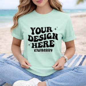 May include: A pale green t-shirt with rolled sleeves displaying the text "YOUR DESIGN HERE" in a bold, black font. Below the text is the word "KIWIBERRY" in a smaller font. The model is wearing blue jeans and white trainers.