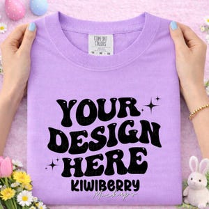 May include: A lavender Comfort Colors t-shirt with the text "YOUR DESIGN HERE" in a retro font. The shirt is held by hands with light blue nail polish. The bottom of the shirt has the text "KIWIBERRY Mockups".