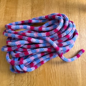 May include: A coiled jump rope with a vibrant pattern of pink, blue, and purple. The rope is made of a thick, braided material. The colors are arranged in a repeating pattern, creating a visually appealing design. The rope is lying on a wooden surface.