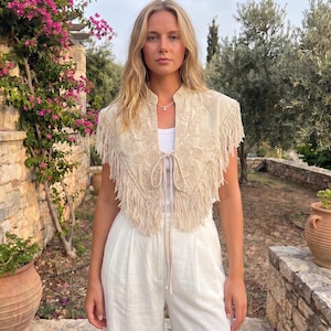 Può includere: Un gilet beige senza maniche con intricati ricami tono su tono e lunghe frange lungo i bordi. Il capo presenta una chiusura frontale a laccetti ed è indossato sopra un semplice top bianco abbinato a pantaloni bianchi.