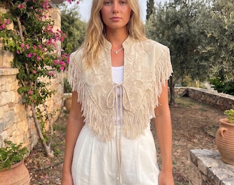 May include: A beige sleeveless vest featuring intricate tonal embroidery and long fringe trim along the edges. The garment has a front tie closure and is worn over a simple white top paired with white trousers, creating a layered look.