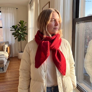Puede incluir: Una bufanda roja brillante sobre una chaqueta acolchada color crema. La bufanda es de un material suave y está envuelta alrededor del cuello. La imagen está tomada en interiores, con luz natural que entra por una ventana.