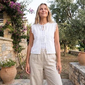 May include: A white sleeveless vest featuring intricate eyelet embroidery and a delicate lace trim along the armholes. The garment has a front tie closure with thin fabric strings and is worn over a simple white top paired with beige linen trousers.